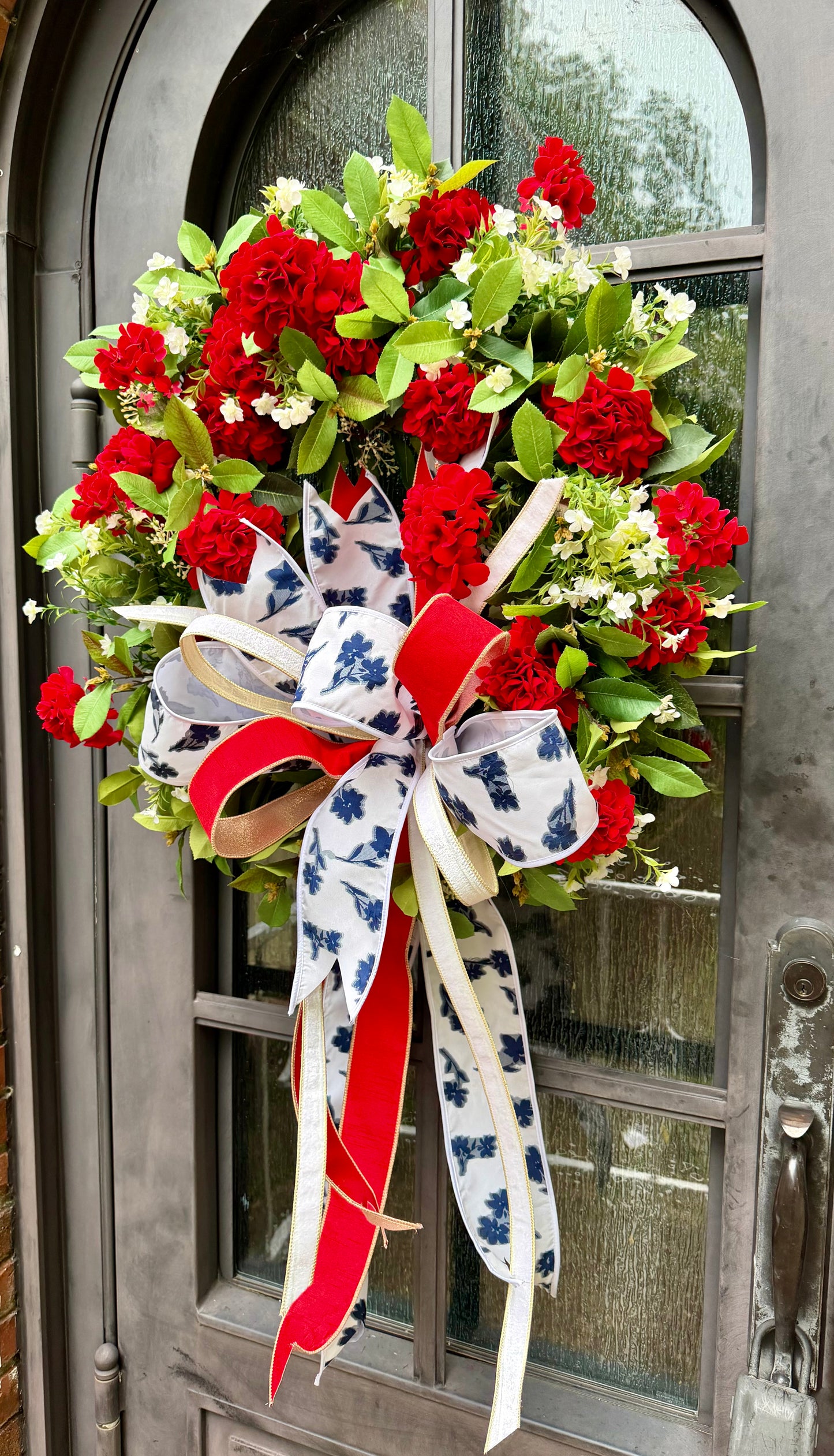 JME handmade faux wreath of red geraniums, white florals, and mixed greenery. Paired with a JME patriotic floral bow! About 27 inches in diameter.