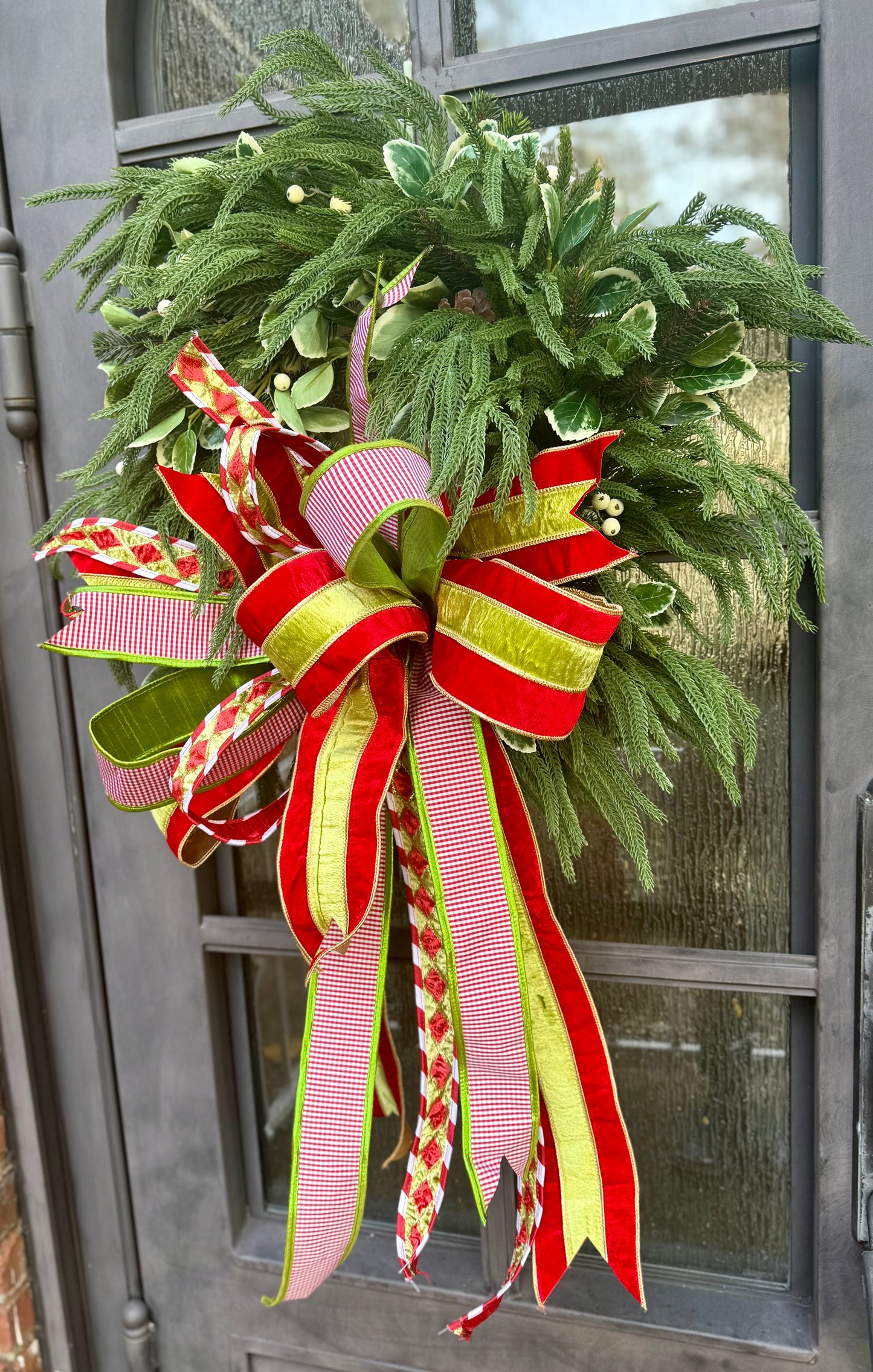 Faux Droopy northfolk wreath. Handmade by JME paired with a JME Handtied green and red with touches of white bow. About 24 inches wide.