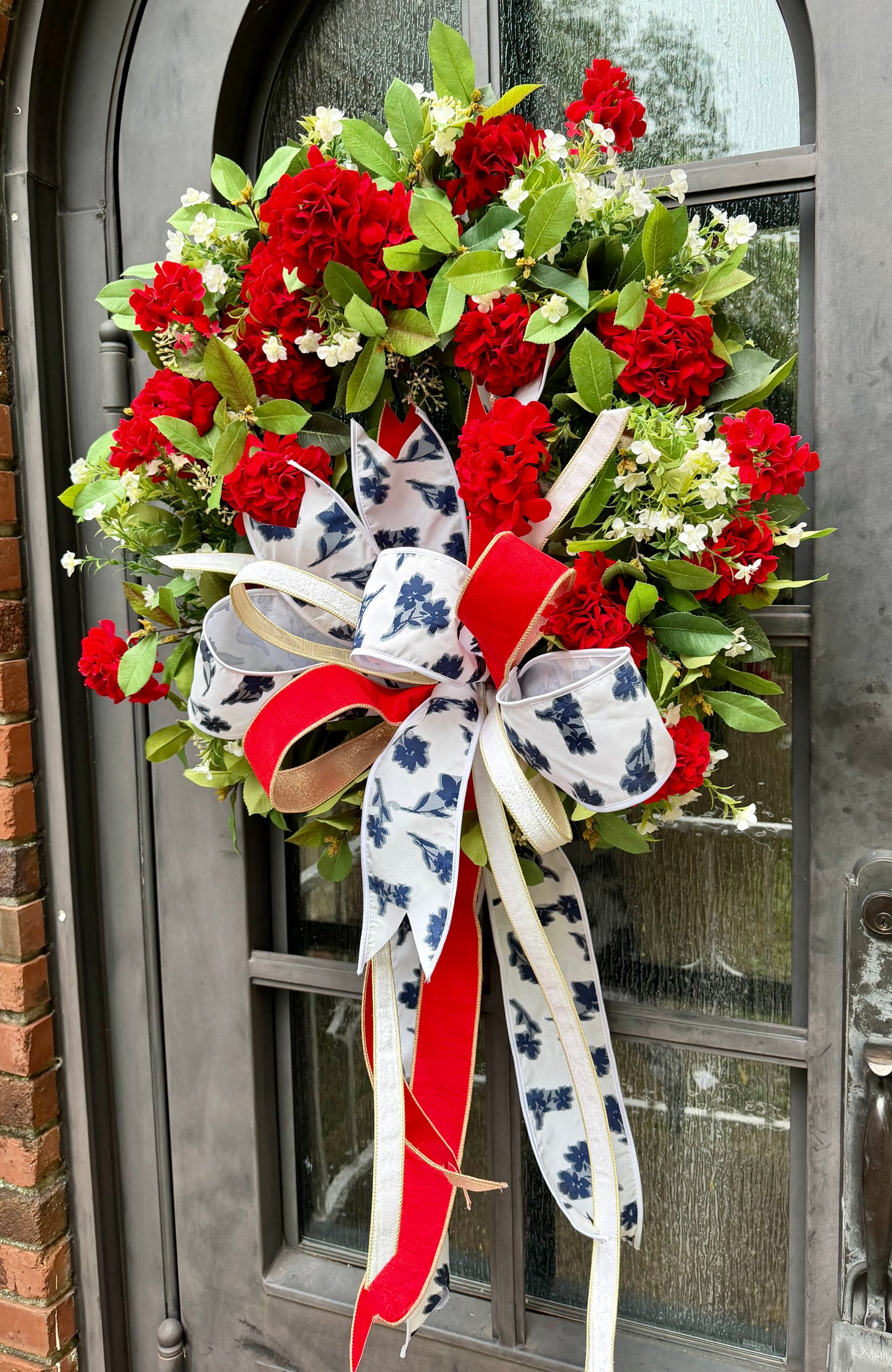 JME handmade faux wreath of red geraniums, white florals, and mixed greenery. Paired with a JME patriotic floral bow! About 27 inches in diameter.