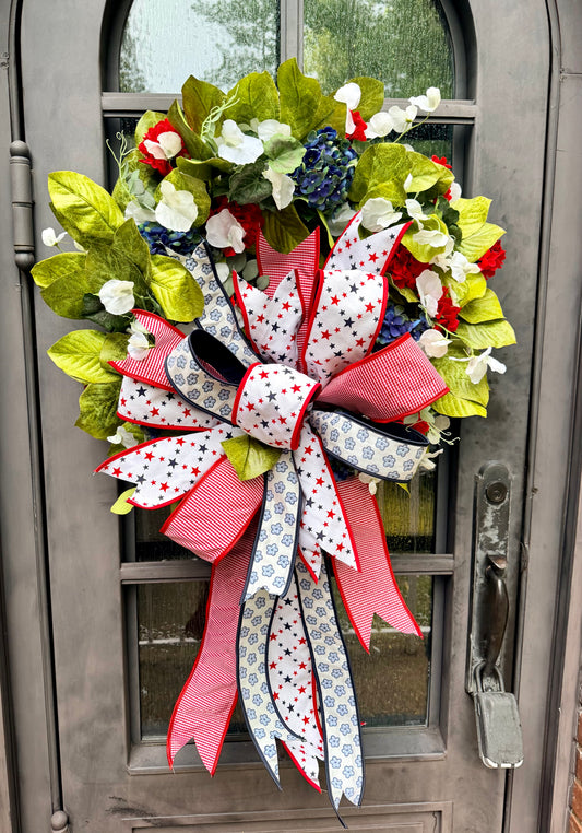 JME handmade faux wreath of magnolias  white sweetpeas, blue hydrangeas, and red geraniums with mixed greenery. Paired with a JME patriotic star bow! About 27 inches in diameter.