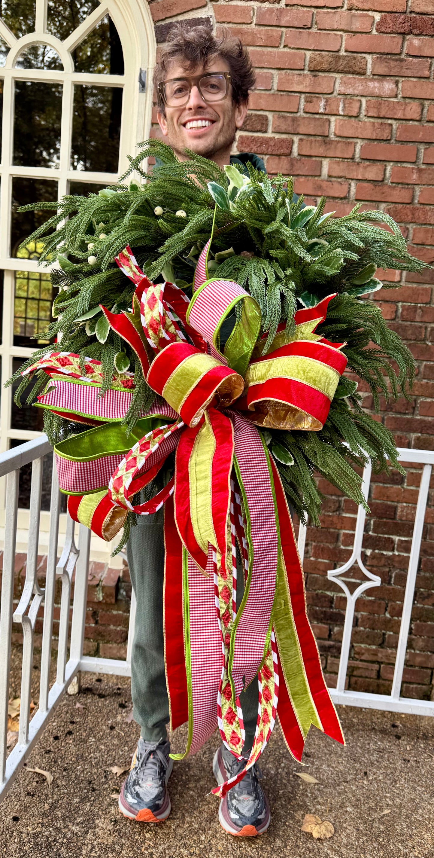 Faux Droopy northfolk wreath. Handmade by JME paired with a JME Handtied green and red with touches of white bow. About 24 inches wide.