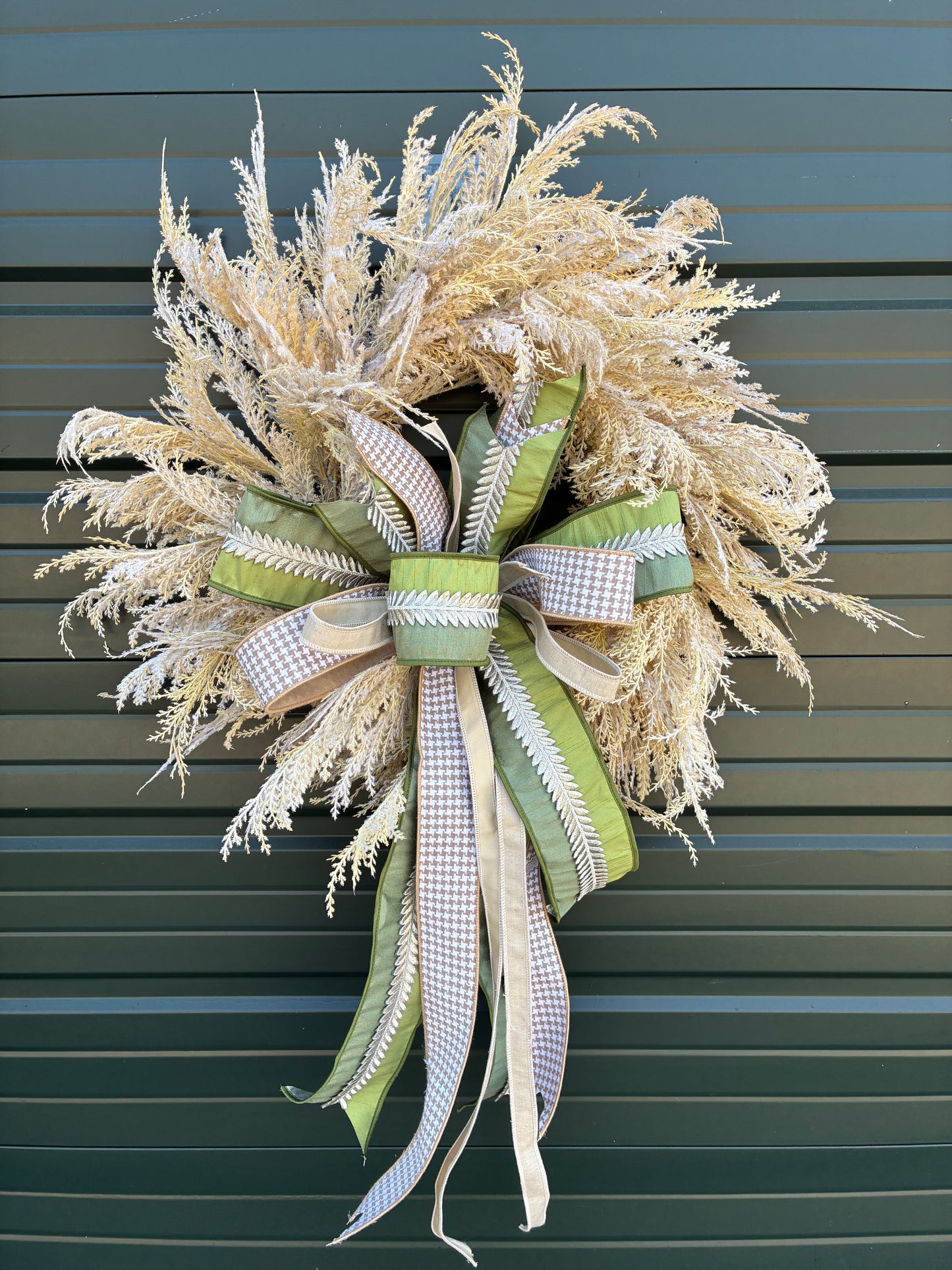 Faux Pampas Grass Wreath paired with a JME hand tied bow featuring a green ribbon with silver leaf embellishment and tan houndstooth ribbon combo. Approximately 28 inches in diameter. Some fluffing may be required upon arrival.