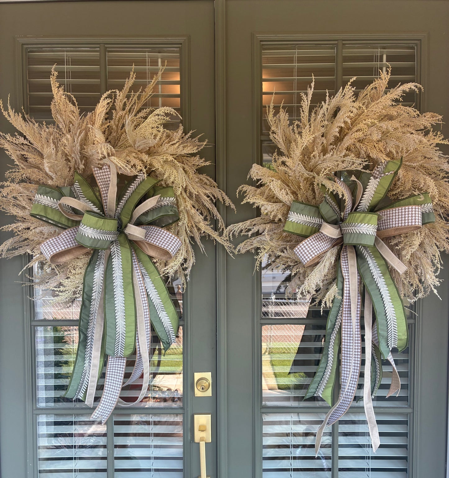 Faux Pampas Grass Wreath paired with a JME hand tied bow featuring a green ribbon with silver leaf embellishment and tan houndstooth ribbon combo. Approximately 28 inches in diameter. Some fluffing may be required upon arrival.