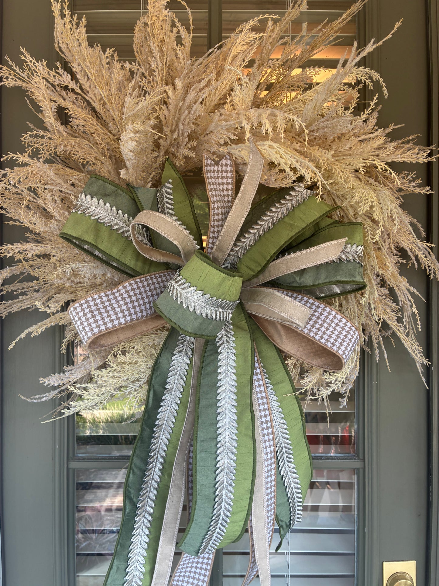 Faux Pampas Grass Wreath paired with a JME hand tied bow featuring a green ribbon with silver leaf embellishment and tan houndstooth ribbon combo. Approximately 28 inches in diameter. Some fluffing may be required upon arrival.